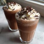 Two tall glasses filled with blended frozen hot chocolate, topped with whipped cream and chocolate shavings.