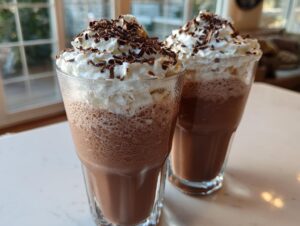 Two tall glasses filled with blended frozen hot chocolate topped with whipped cream and chocolate shavings.