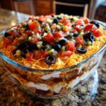 A close-up view of a vibrant 7 layer dip showcasing layers of cream cheese, beans, and topped with shredded cheese, olives, and pico de gallo.