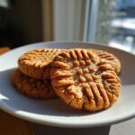 Three golden brown peanut butter cookies with classic crosshatch fork marks stacked on a white plate.
