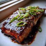 Close-up of a perfectly cooked piece of teriyaki salmon covered in a thick, glossy glaze, topped with sesame seeds and sliced green onions.