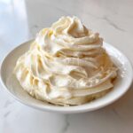 A generous swirl of light, creamy vanilla buttercream frosting piled high in a white bowl.
