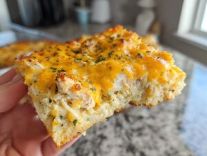 A hand holding a thick, square slice of cheesy chicken and rice casserole topped with melted cheddar and parsley.