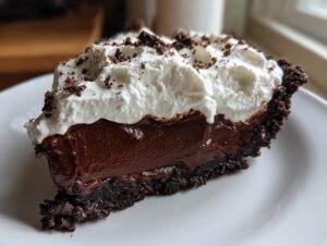 A close-up of a rich slice of Christmas chocolate cream pie featuring a dark crust, thick chocolate filling, and whipped cream topping.