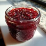 Close-up of a small glass jar filled with glistening, deep red homemade christmas jam, catching the sunlight.