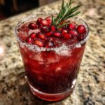 A close-up of a vibrant red cranberry pomegranate margarita in a sugar-rimmed glass, topped with pomegranate seeds and a rosemary sprig.
