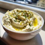 A close-up of creamy dirty martini dip in a white bowl, garnished with chopped green olives, herbs, and olive oil.