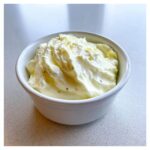 A white bowl filled with thick, creamy, pale yellow garlic aioli, showing swirls and flecks of herbs.