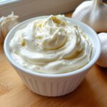 A bowl of thick, creamy garlic sauce, garnished with spices, next to whole garlic bulbs.