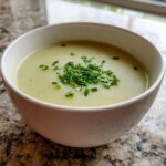 A close-up of creamy, pale green leek soup in a white bowl, garnished with fresh chopped chives.
