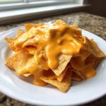 Close-up of tortilla chips smothered in thick, bright orange nacho cheese sauce on a white plate.