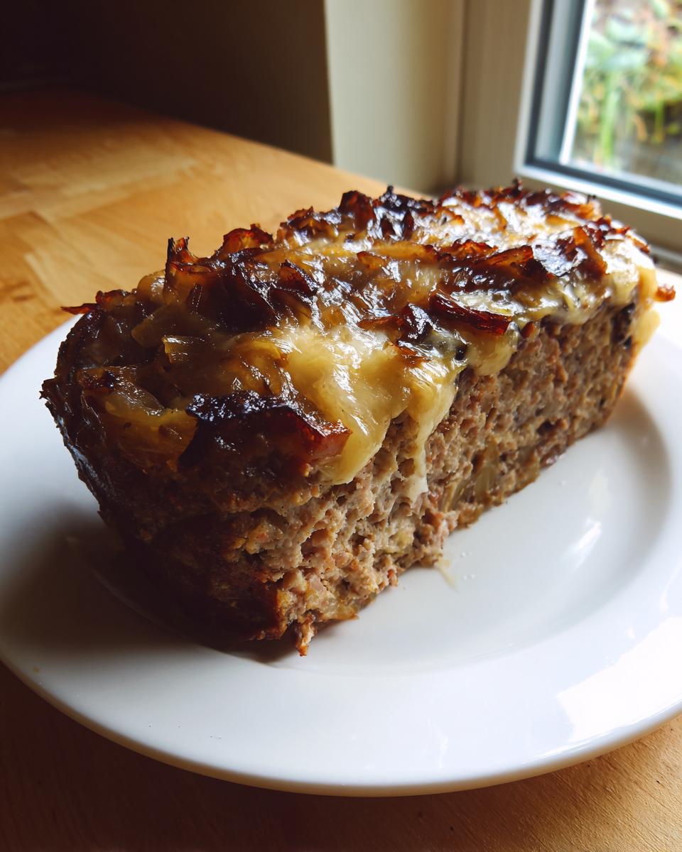 A thick slice of crockpot french onion meatloaf topped with melted cheese and caramelized onions on a white plate.