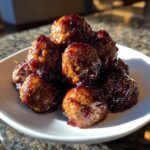 A close-up pile of glistening Crockpot grape jelly meatballs coated in a dark, sweet sauce on a white plate.