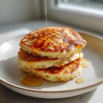 Close-up of a stack of three fluffy banana pancakes being drizzled with golden syrup.