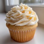 A single, fluffy vanilla cupcake topped with a generous swirl of creamy, pale yellow vanilla frosting.