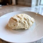 A ball of pale, slightly textured homemade marzipan recipe dough resting on a white plate.