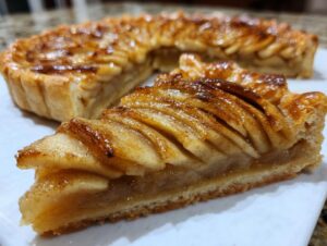 A close-up slice of a beautiful, glazed apple tart showing thinly sliced, caramelized apples, with the rest of the apple tart blurred in the background.