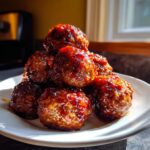 A close-up of several glazed cranberry meatballs piled high on a white plate, glistening under bright light.