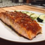 A perfectly cooked fillet of salmon covered in a thick, glossy, amber glaze, served with sliced cucumbers.