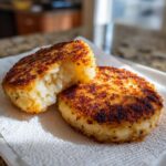 Two crispy, golden brown mashed potato cakes resting on a paper towel, with one cut open showing the fluffy interior.