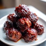 Close-up of a stack of glistening, saucy grape jelly meatballs piled high on a white appetizer plate.