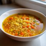 Close-up of a white bowl filled with vibrant italian penicillin soup, featuring small pasta pearls and diced carrots in a rich broth.