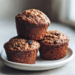 A stack of three dark, moist bran muffins topped with visible raisins, resting on a small white plate.