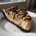 A decadent slice of peanut butter pie with a dark crust, topped with peanut butter drizzle, chocolate, and crushed Oreos.