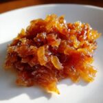 A close-up mound of deeply golden, glossy caramelized onions on a white plate, showing rich color.