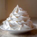 A tall, perfectly piped swirl of white frosting, resembling cream cheese frosting, sits on a small white plate.