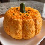 A festive, pumpkin-shaped pumpkin cheese ball covered in shredded cheddar cheese and topped with a green stem.