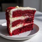 A tall, moist slice of red velvet cake layered with thick white cream cheese frosting, sitting on a white plate.