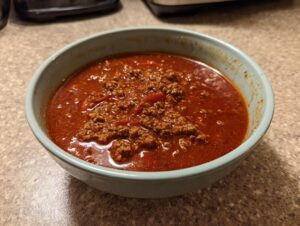 A close-up of a light blue bowl filled with rich, thick crockpot chili featuring ground beef in a deep red sauce.