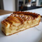 A close-up of a thick slice of baked apple tart showing layered, caramelized apple slices on a buttery crust.