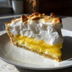 A tall slice of lemon meringue pie showing bright yellow curd and fluffy, toasted meringue.