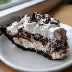 A decadent slice of ice cream pie featuring layers of chocolate crust, vanilla filling, fudge swirls, and whipped cream topping.