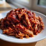 A close-up of shredded, saucy Slow Cooker Pulled Pork piled high on a white plate, glistening under natural light.