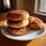 Three soft Snickerdoodle cookies stacked on a white plate next to one cookie, coated in cinnamon sugar.