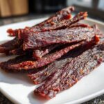 A pile of dark, richly seasoned venison jerky strips sprinkled with sesame seeds and spices, resting on a white plate.