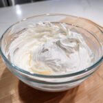 A clear glass bowl filled with perfectly whipped, bright white royal icing recipe mixture showing stiff peaks.