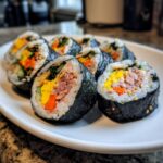 Several slices of homemade tuna kimbap featuring tuna filling, yellow egg, orange carrots, and green vegetables, sprinkled with sesame seeds.