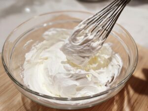 Close-up of thick, white, fluffy mixture being whisked in a clear glass bowl, essential for a royal icing recipe.