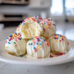 A stack of white chocolate coated sugar cookie truffles topped with rainbow sprinkles, one is bitten open.