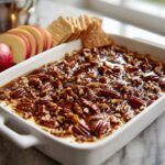 A white baking dish filled with creamy pecan pie dip topped with a glossy caramel pecan mixture, served with apple slices and crackers.