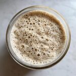 Overhead view of a highly active sourdough starter in a glass jar, covered in small, uniform bubbles.