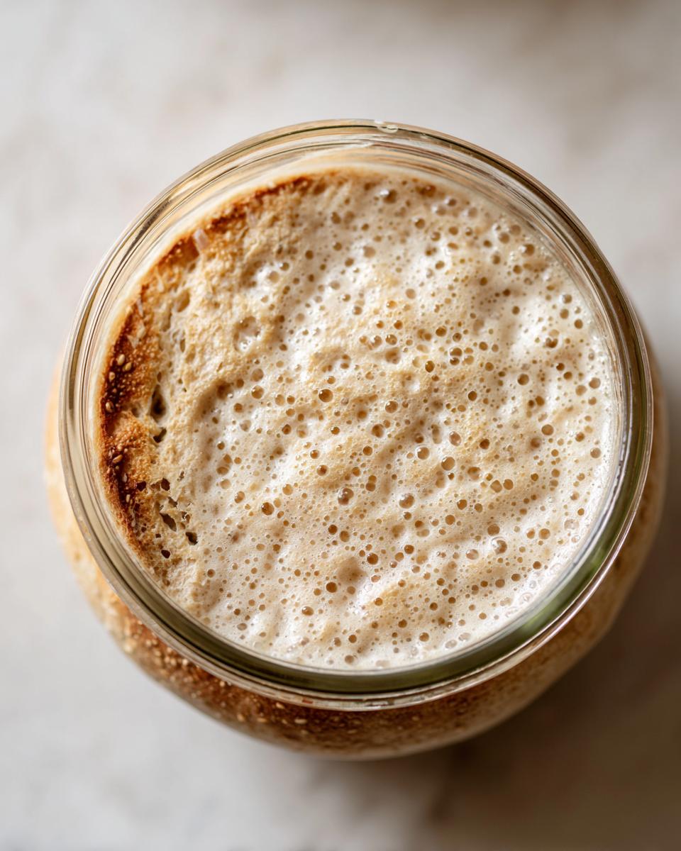 Overhead view of a very active sourdough starter in a glass jar, showing a thick layer of foam and many bubbles.