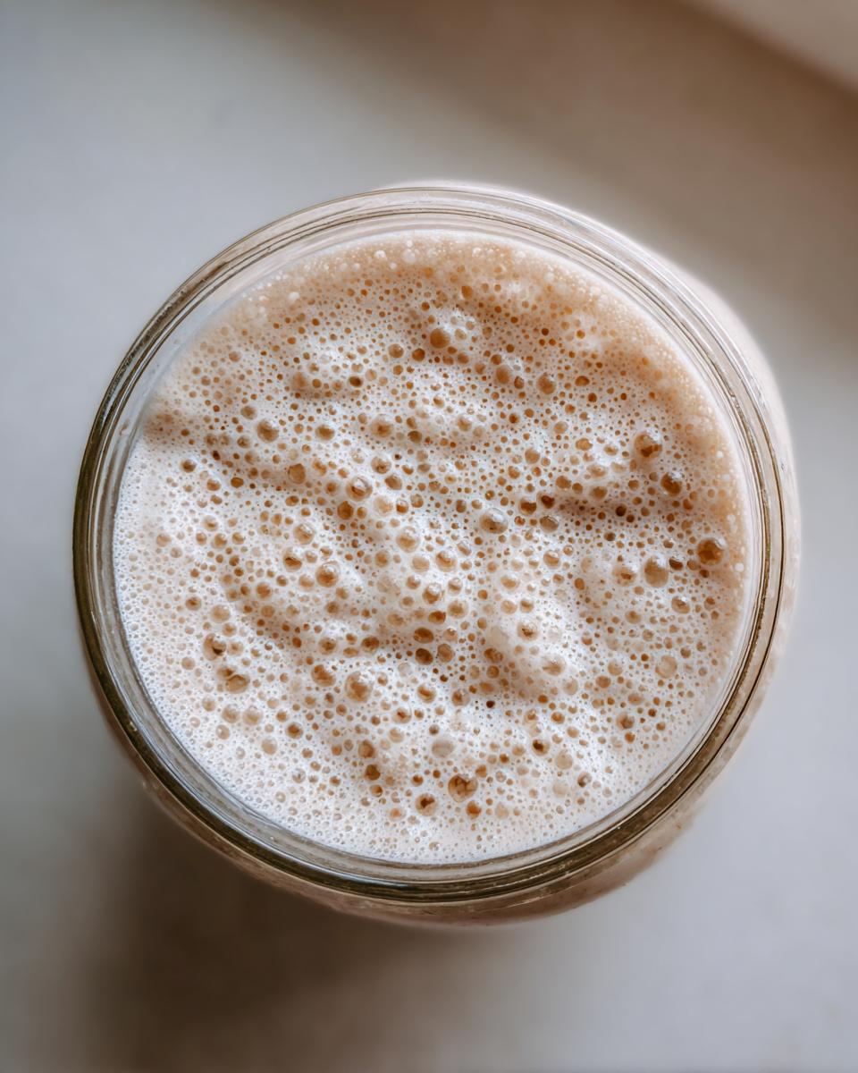 Overhead view of an active sourdough starter in a glass jar, showing a thick layer of small, active bubbles.
