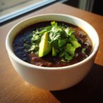A close-up of rich, dark black bean soup in a white bowl, garnished with fresh avocado slices and cilantro.