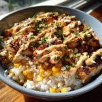 Close-up of a flavorful chicken rice bowl topped with seasoned chicken, corn, white cheese, and a creamy sauce.