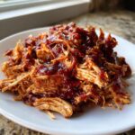 Close-up of tender, shredded pulled chicken generously coated in rich, dark BBQ sauce on a white plate.